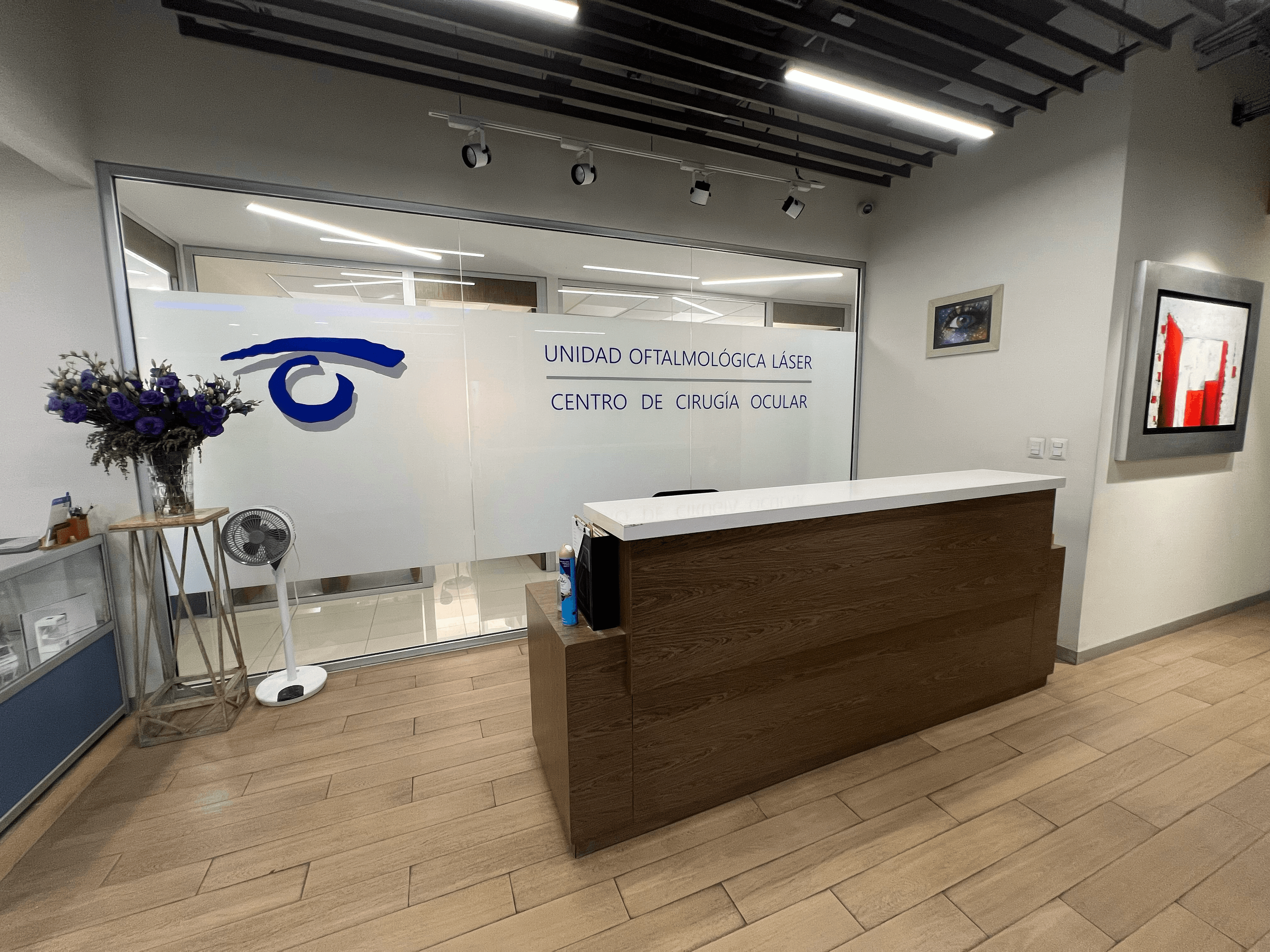 Recepción de Unidad Oftalmológica Láser con mostrador de madera, flores moradas y pared de vidrio.