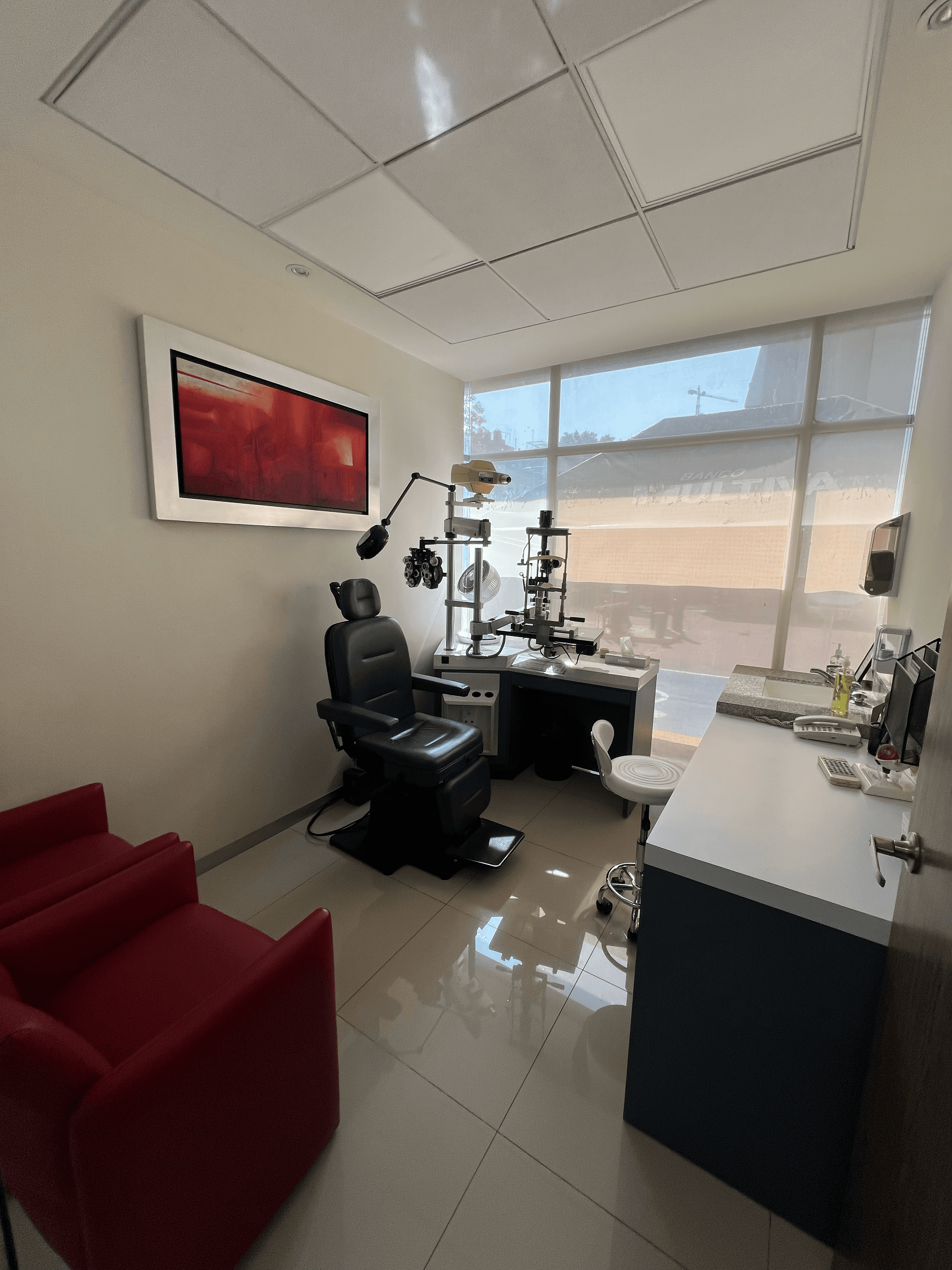 Consultorio de optometría con silla de examen negra, equipo especializado, ventana grande y sillones rojos.