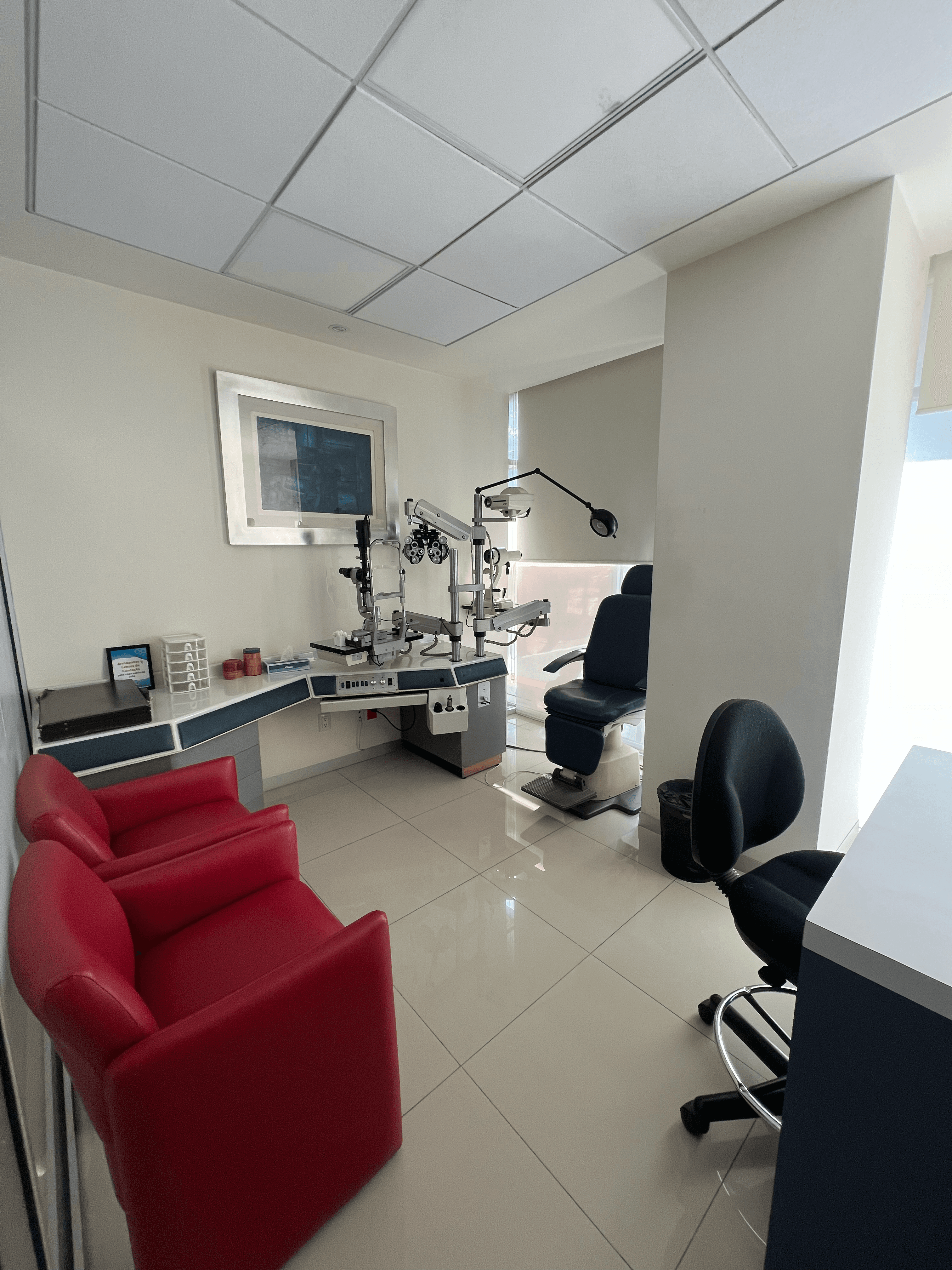 Consultorio oftalmológico equipado con foróptero, silla de examen azul y dos sillones rojos de cuero.