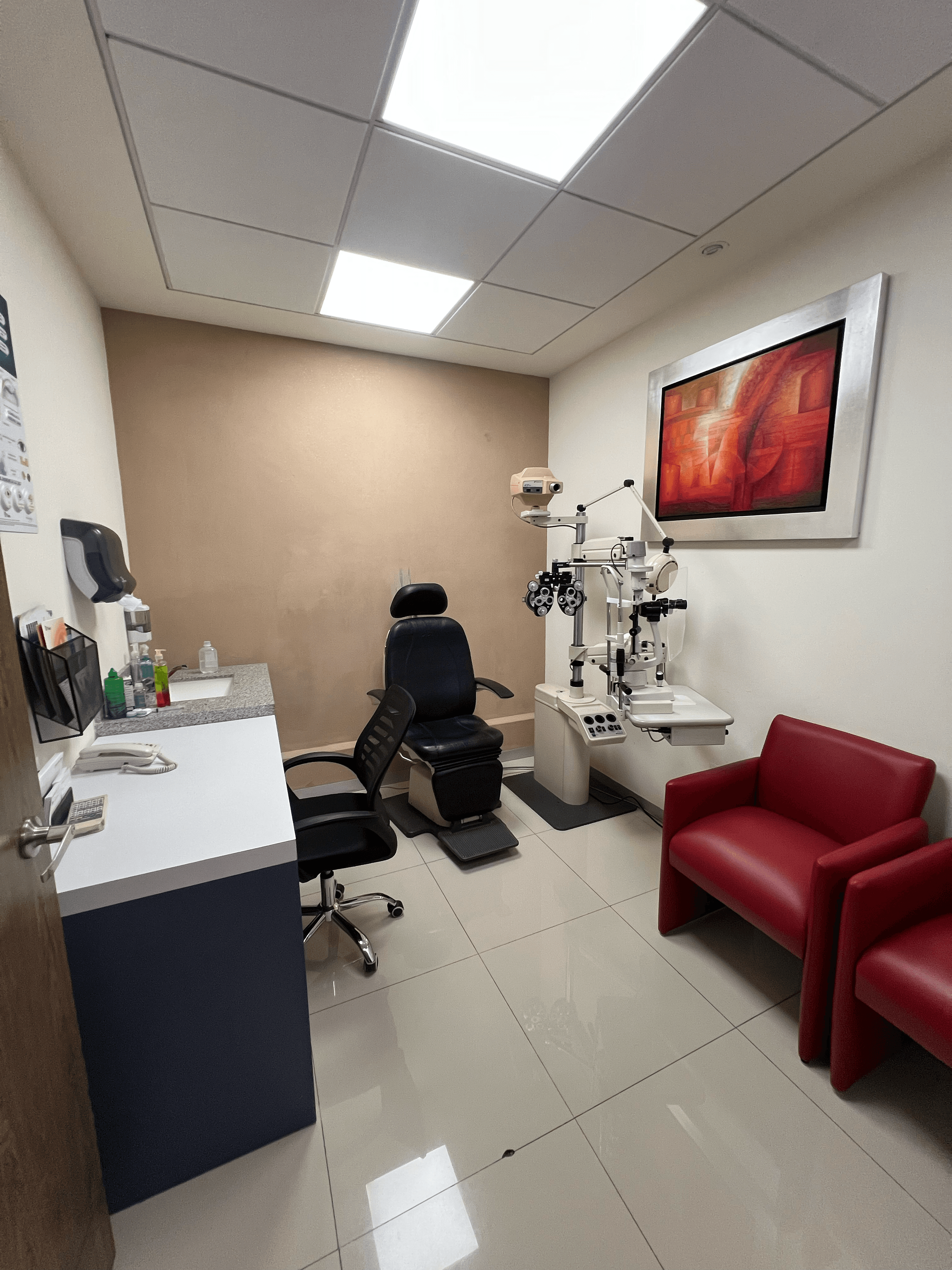 Consultorio de oftalmología con equipo de examen, silla negra, escritorio blanco y sillones rojos.